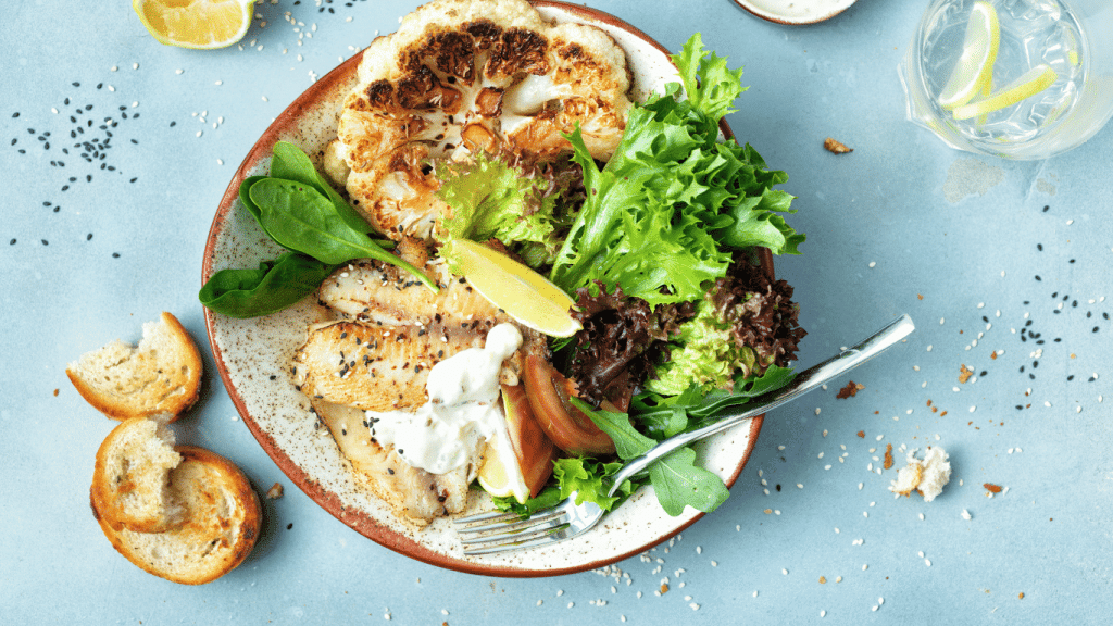 A delectable plate of grilled fish and cauliflower steaks accompanied by a fresh mixed greens salad is presented, perfectly embodying the nutritious and flavorful meals you can create with Chefs Plate. The plate is garnished with lemon wedges, a dollop of creamy sauce, and toasted bread slices on the side. Sesame seeds are sprinkled over the golden-brown fish, adding a subtle crunch. This inviting dish is a testament to the kind of culinary delights you can enjoy at a discount with the Chefs Plate coupon code, making gourmet home-cooking both accessible and affordable.