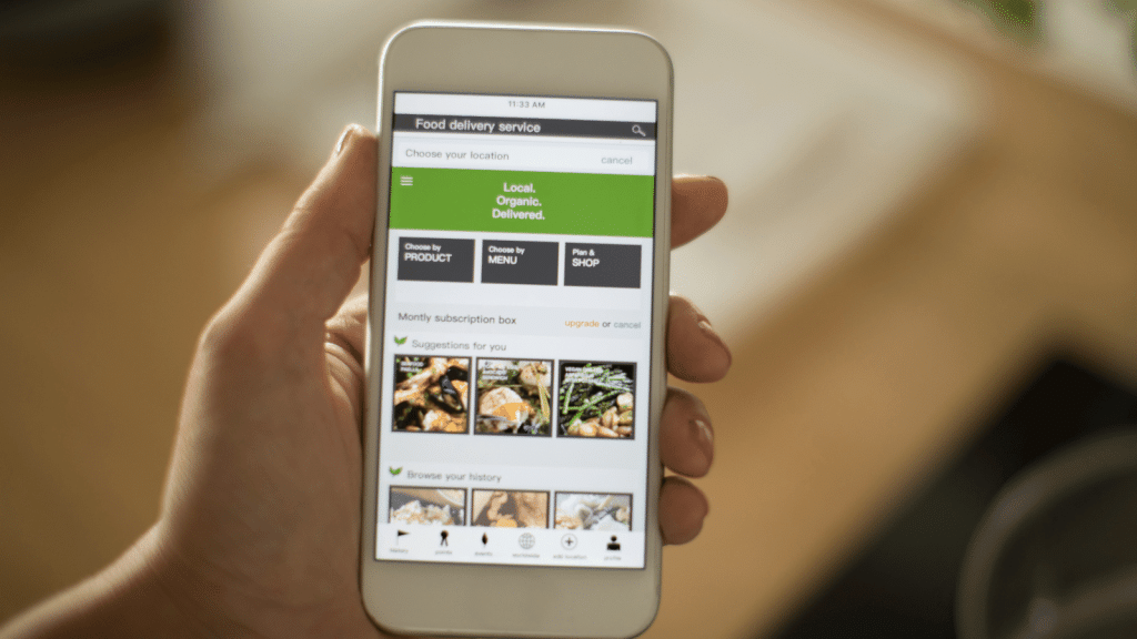 This image shows a person's hand holding a smartphone displaying a food delivery service application. The screen highlights options for local, organic meal deliveries with a menu for product choice, menu selection, and shopping plans. It suggests a monthly subscription box and offers tailored suggestions based on user history, emphasizing the personalized and user-friendly experience one should consider when choosing the best prepared meal delivery service.