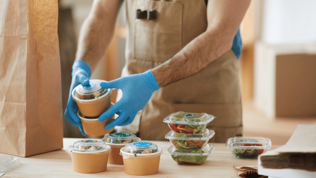 This image features a person in a brown apron and blue gloves, carefully packing a variety of prepared meals into a paper bag. The meals include clear containers with green salads and round paper bowls with lids, indicating different meal options available for customization in a prepared meal delivery service. The setting suggests a professional kitchen or packing area, emphasizing the service’s commitment to hygiene and customer personalization.