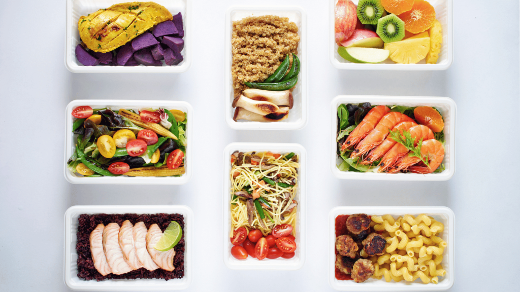 This image presents an array of diverse and colourful prepared meals arranged neatly in white containers, showcasing the variety one can enjoy when maximizing the benefits of a prepared meal delivery service. The dishes include a chicken breast with purple potatoes, a mixed vegetable salad with cherry tomatoes, quinoa with green veggies, a vibrant fruit assortment, fresh prawns on a bed of greens, a serving of sliced turkey over black rice with lime, stir-fried vegetables with noodles, and meatballs with pasta. This selection exemplifies the convenience, nutritional balance, and culinary variety that such services offer.