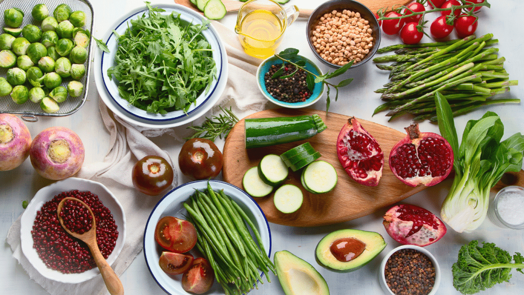 Image of a vibrant array of fresh produce and legumes spread out on a white surface, ideal for a nutritious meal plan for a week. Ingredients include Brussels sprouts, arugula, chickpeas, black peppercorns, tomatoes on the vine, asparagus, and a halved pomegranate. Also featured are sliced zucchini on a wooden cutting board, an open avocado with a pit, and leafy greens like bok choy and kale. These colorful, wholesome foods are perfect for creating healthy recipes.