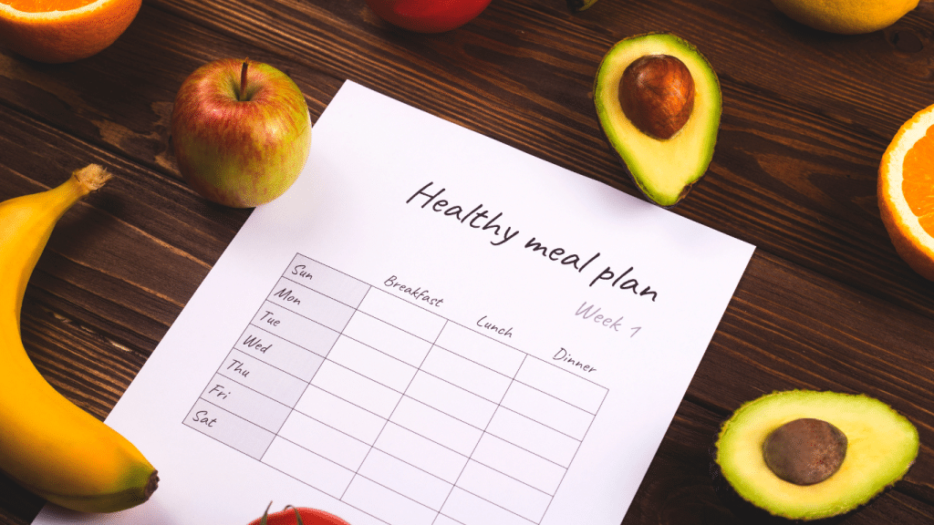 Image of a 'Healthy meal plan' schedule for week 1 on a white paper, surrounded by a variety of fresh fruits like bananas, apples, avocados, and oranges, all on a dark wooden table. The meal plan is blank, ready to be filled with nutritious meal choices for breakfast, lunch, and dinner, embodying a balanced approach to weekly food preparation.