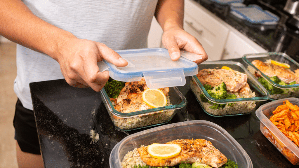 Image showcasing a person's hands securing the lid on a glass meal prep container, which is one of several lined up on a kitchen countertop. Each container is filled with a nutritious meal consisting of grilled chicken, a slice of lemon, broccoli, and a bed of rice. In the foreground, another container holds a tomato-based pasta dish, indicating a diverse meal plan for the week.